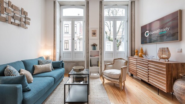 Een moderne woonkamer met een blauwe bank, een glazen salontafel, een houten kast en een groot raam met uitzicht op gebouwen.