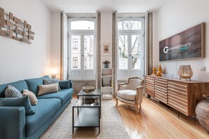 A modern living room with a blue sofa, a glass coffee table, a wooden cabinet, and a large window with a view of buildings. - GuestReady - Chakra Duplex (Porto)