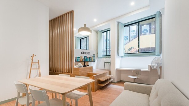 A modern, well-lit room with a wooden dining table, white chairs, a sofa, and a large window with a view of buildings. - GuestReady - A modern stay in Downtown (Porto)
