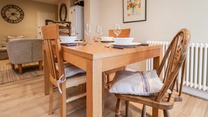 Een eethoek met een houten tafel voor vier personen, omringd door houten stoelen met kussens. Een witte radiator is aan de rechterkant zichtbaar, en boven de tafel hangt een lijst met een afbeelding.