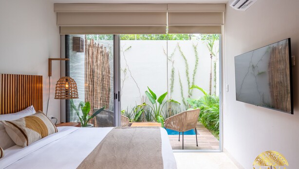 A bedroom with a large bed, a TV mounted on the wall, and a sliding glass door leading to a balcony with plants. - NHOA 108B Lux 1BR Condo Plunge Pool in Aldea Zama (Tulum)