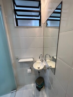 Uma casa de banho moderna com azulejos brancos, uma pia, um espelho, um porta-toalhas e uma pequena lixeira.