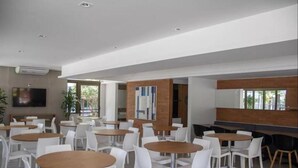A modern dining area with round tables and white chairs, a flat-screen TV mounted on the wall, and a brick accent wall. - LIBERTY PLACE - PARQUE DAS ROSAS (Rio de Janeiro)
