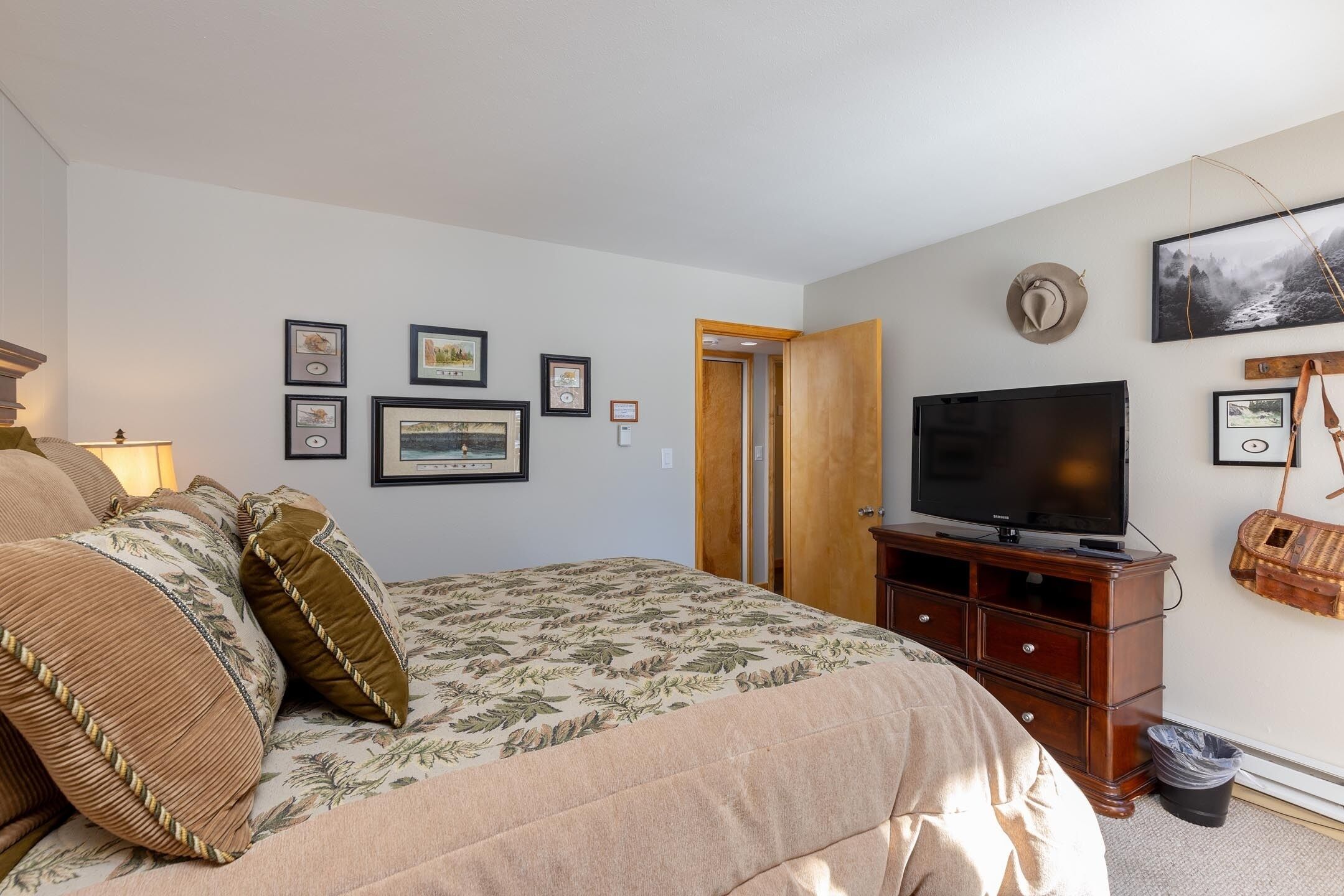 Une chambre à coucher avec un lit, un téléviseur posé sur un meuble en bois, des tableaux encadrés accrochés au mur et une porte donnant sur une autre pièce.
