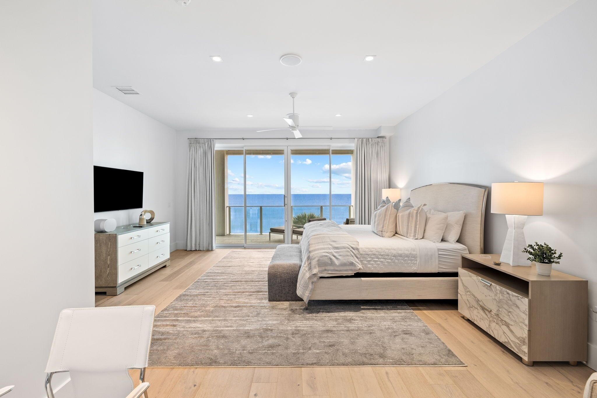 A modern bedroom with a large bed, a TV, a desk, and a view of the sea.