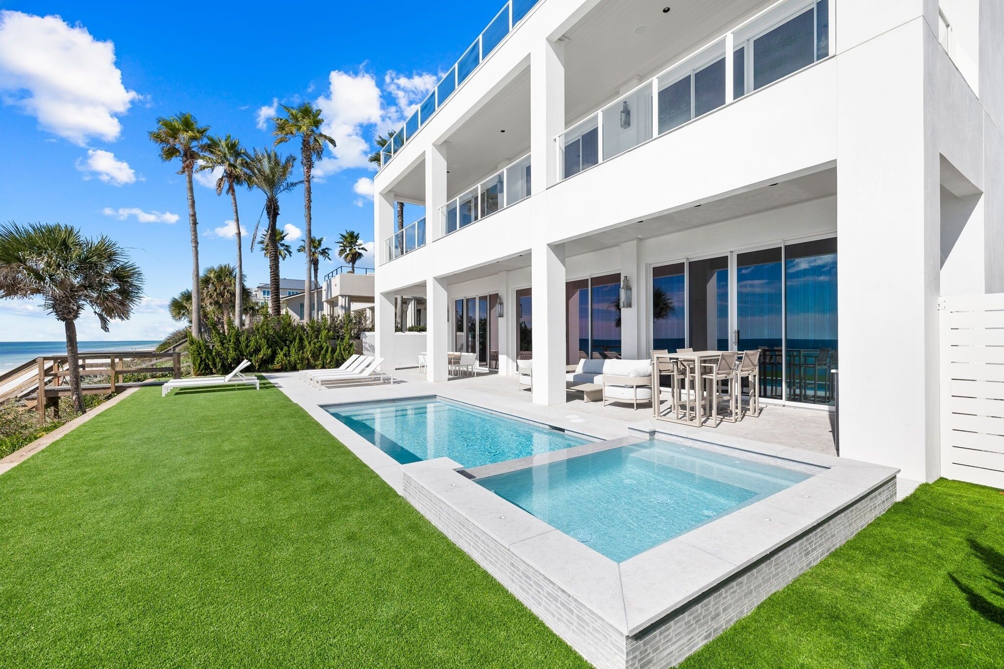 Una casa moderna de dos pisos con piscina, mobiliario exterior y vista al océano.