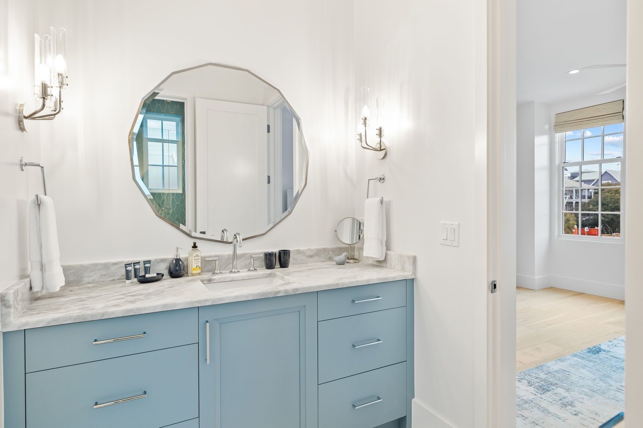 A bathroom with a large oval mirror, a marble countertop, and light blue cabinets.