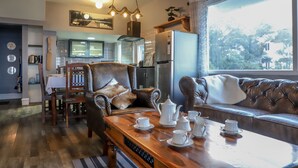 A cozy living room with a wooden coffee table set with tea cups and a teapot, leather sofas, a dining area with a wooden table and chairs, and a kitchen in the background. - Eagles Wings Abode (Coonoor)