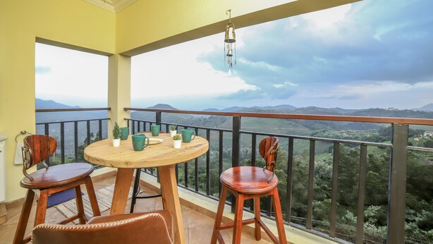 A balcony with a wooden table and chairs, a hanging lantern, and a scenic view of mountains and greenery. - Eagles Wings Abode (Coonoor)