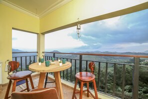A balcony with a wooden table and chairs, a hanging lantern, and a scenic view of mountains and greenery. - Eagles Wings Abode (Coonoor)