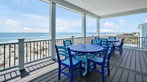 Uma área externa coberta com deck de madeira, cadeiras azuis e uma mesa, com vista para uma praia, o oceano e edifícios ao longe.