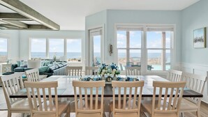 Uma área de jantar com vista para o oceano, com uma mesa, cadeiras, um sofá e um arranjo de flores.
