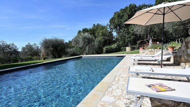 Ein Swimmingpool mit klarem, blauem Wasser, umgeben von Steinmauern und einer Steindeck. Liegestühle und ein großer Sonnenschirm spenden Schatten.