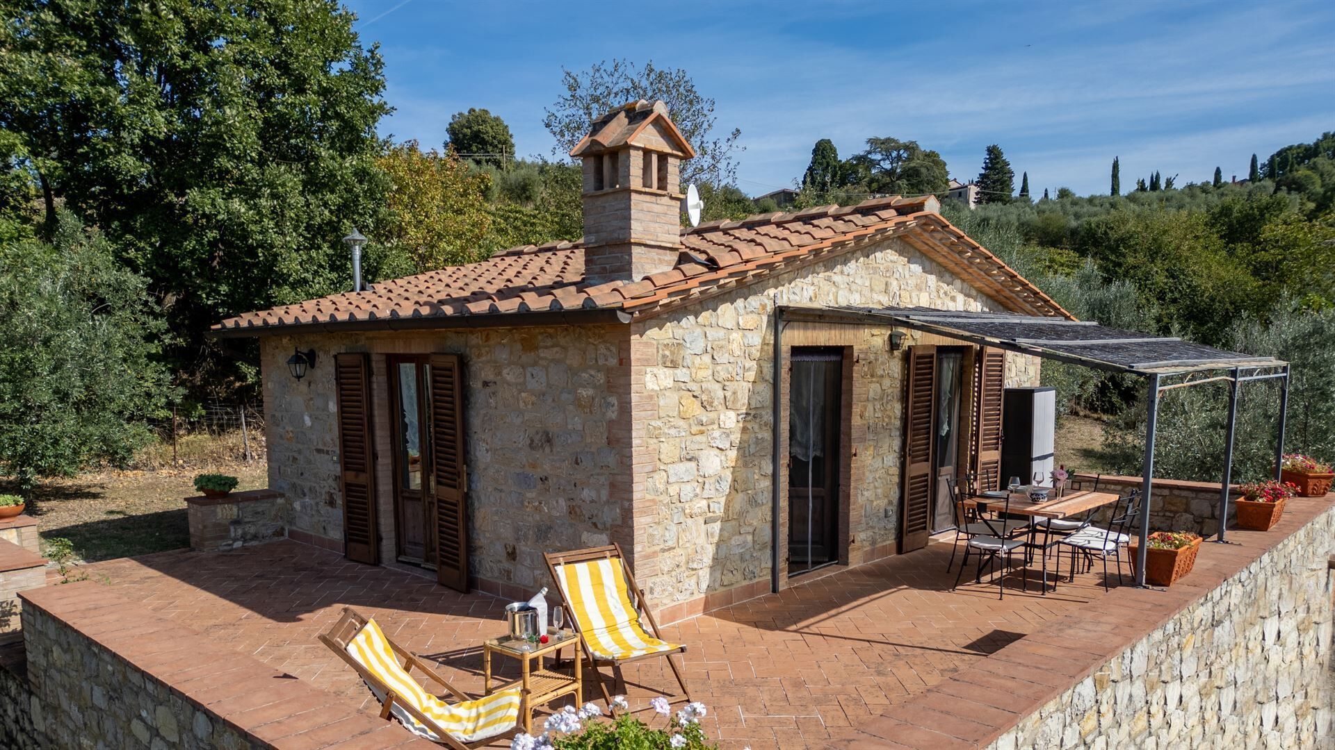 A stone house with a tiled roof, a covered patio with a table and chairs, and a garden area with potted plants.