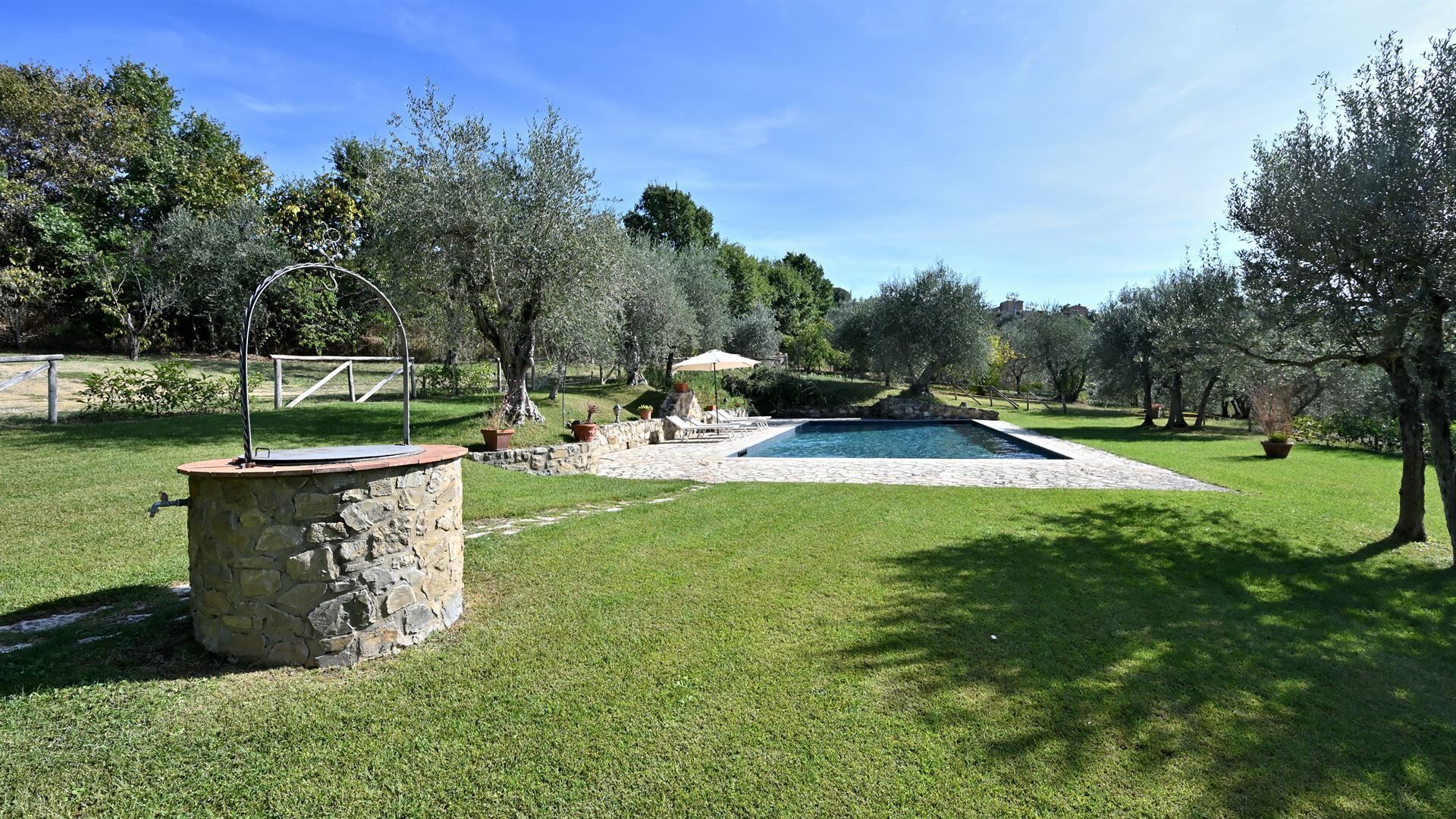 A stone well with a faucet, a swimming pool, and a stone wall in a grassy yard with trees.