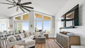 Uma sala de estar espaçosa com um sofá grande, um ventilador de teto e vista para a praia.