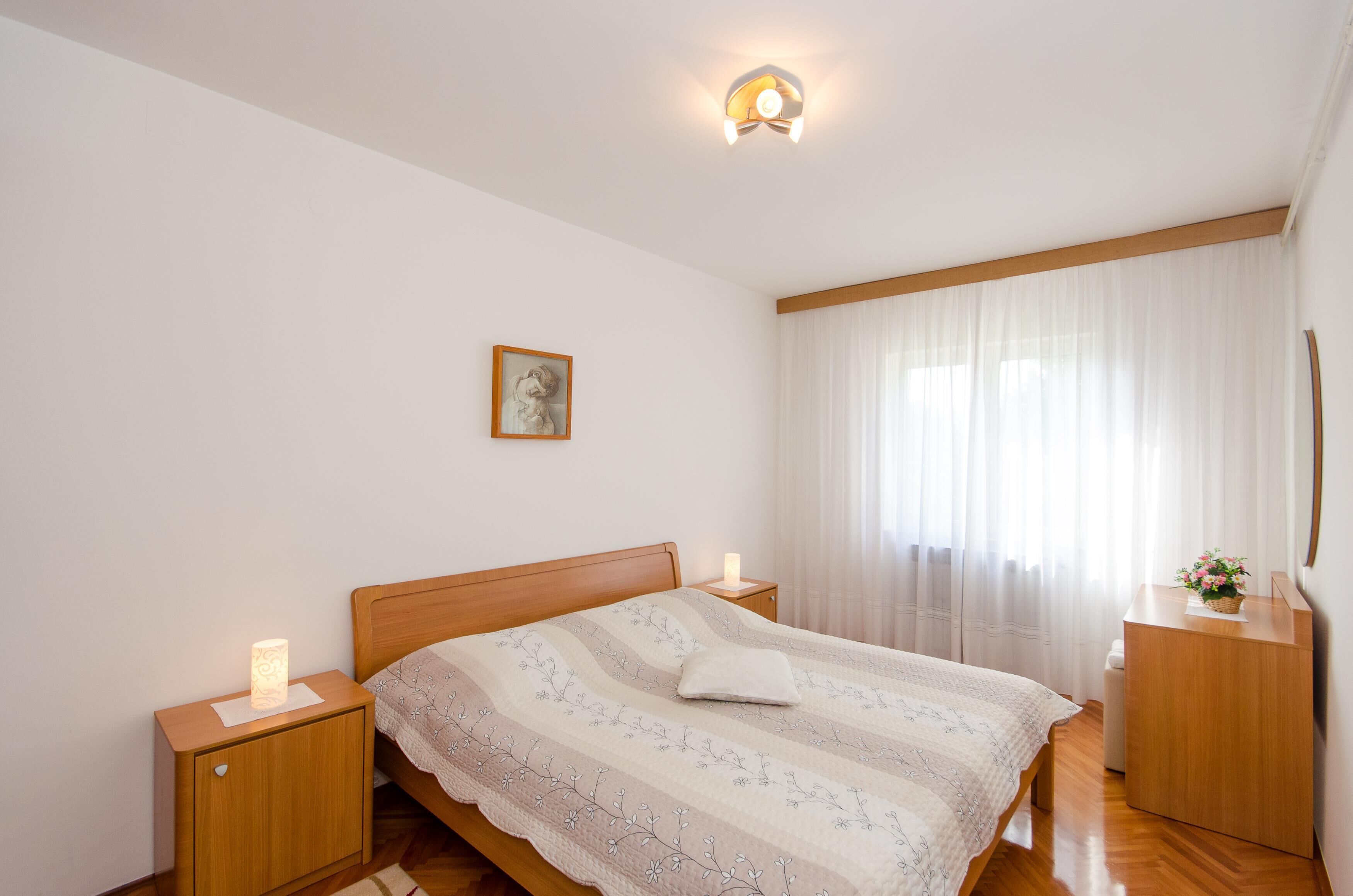 A bedroom with a wooden bed, nightstands, a bedside lamp, a framed picture on the wall, and a window with sheer curtains.