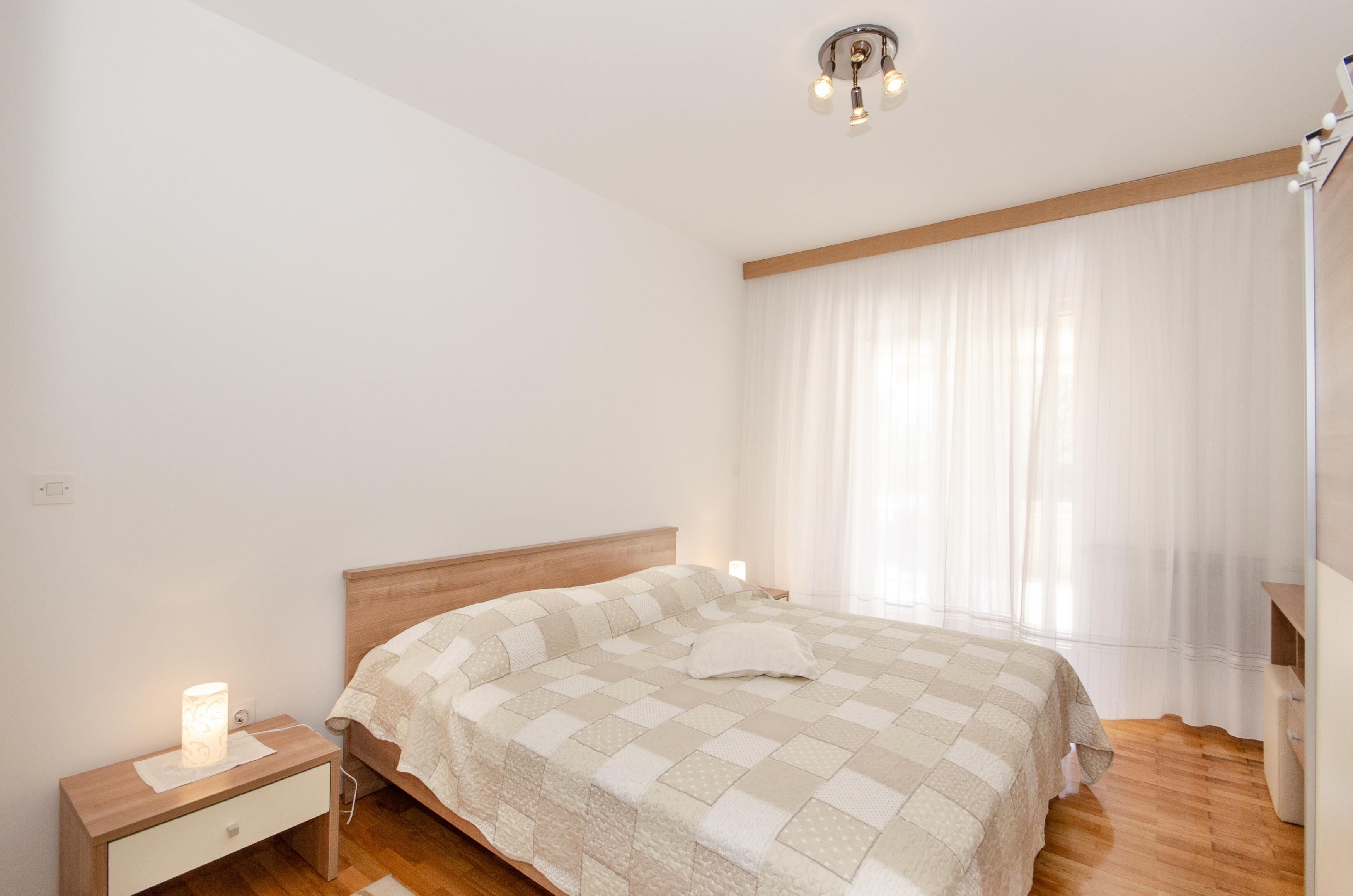 A bedroom with a wooden bed, a bedside table with a lamp, and a window with sheer curtains.