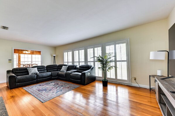 A living room with a sectional sofa, a large window with blinds, a potted plant, and a wooden floor.