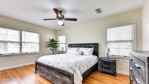 A bedroom with a wooden bed, a nightstand, a dresser, a ceiling fan, and large windows. - Jacksonville Vacation Home w/ Trout River Access (Jacksonville)