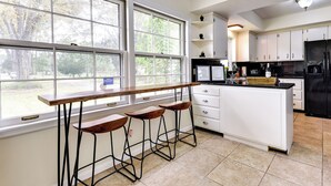 A modern kitchen with a large window, bar stools, white cabinets, and a black refrigerator. - Jacksonville Vacation Home w/ Trout River Access (Jacksonville)