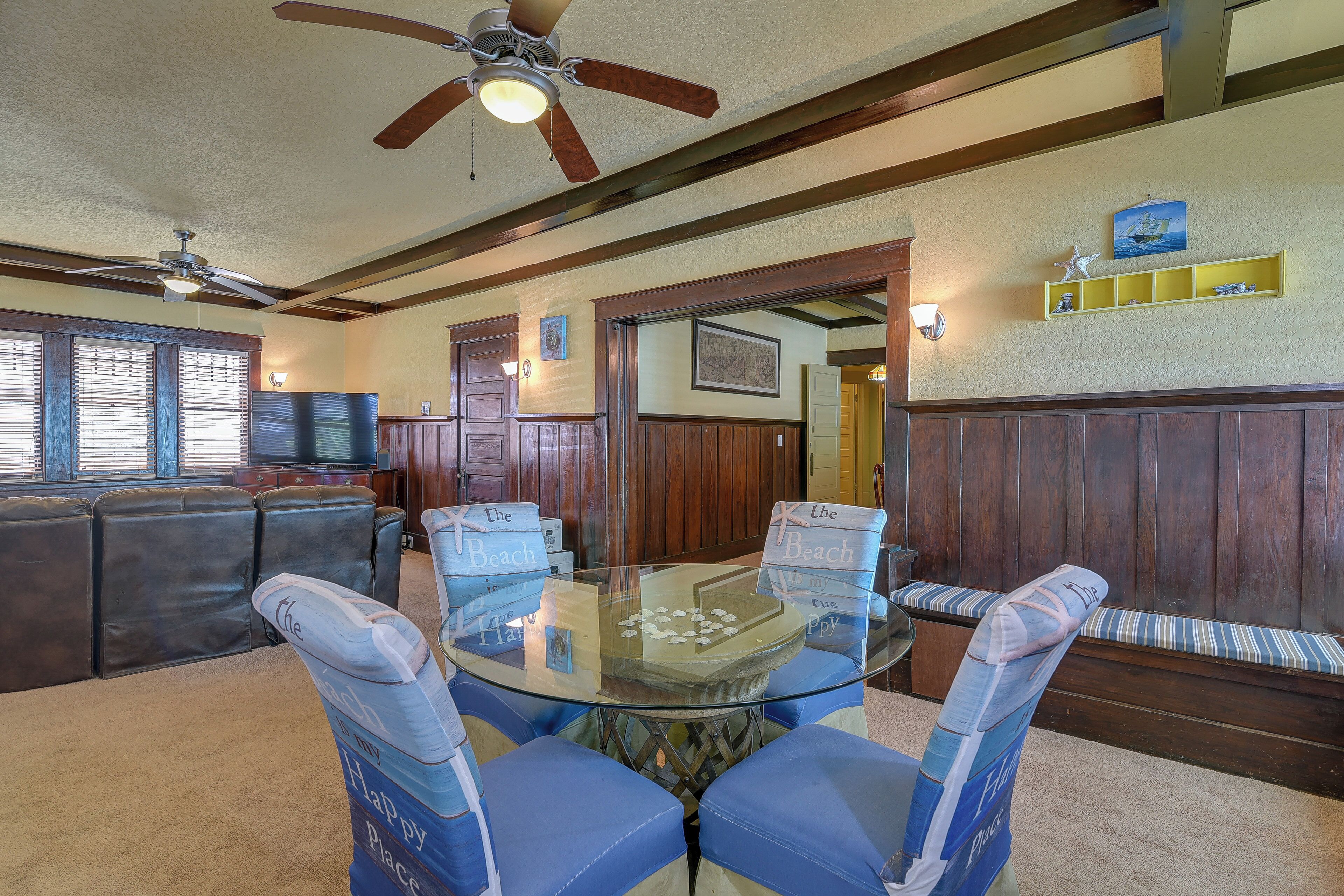 A dining area with a glass-top table surrounded by blue chairs, wooden paneling on the walls, and a ceiling fan.