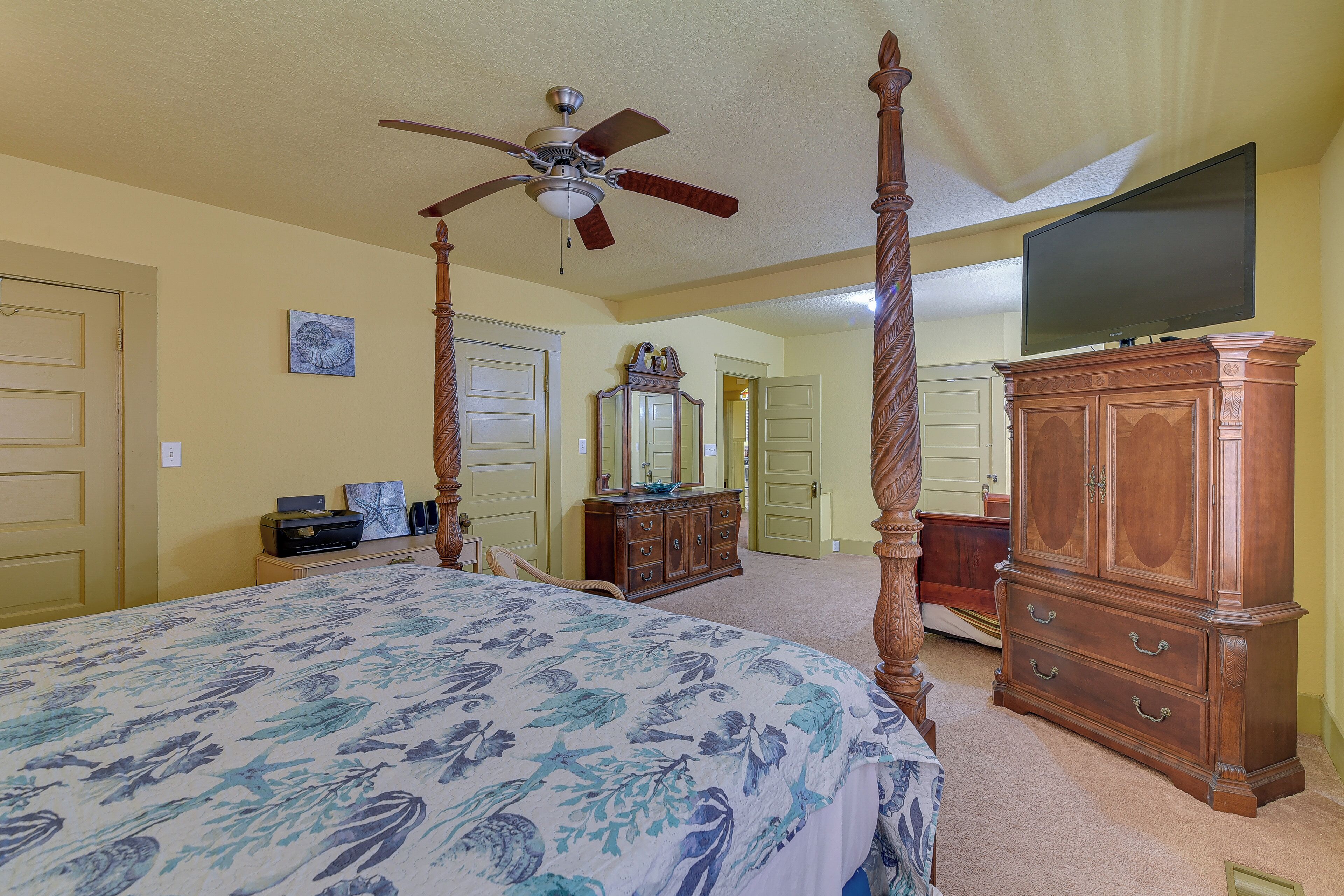 A bedroom with a four-poster bed, a ceiling fan, a dresser, a television, and a door leading to another room.