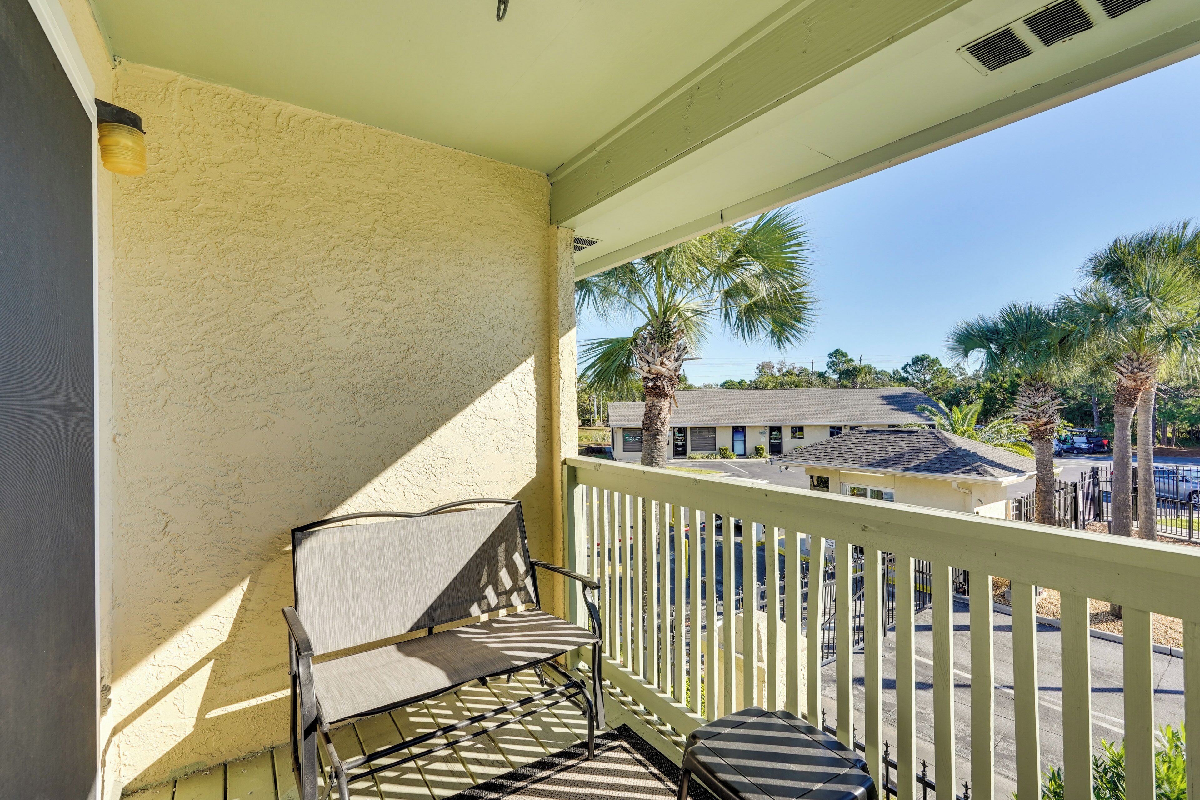 A balcony with a chair and a small table, overlooking a street with palm trees and buildings.