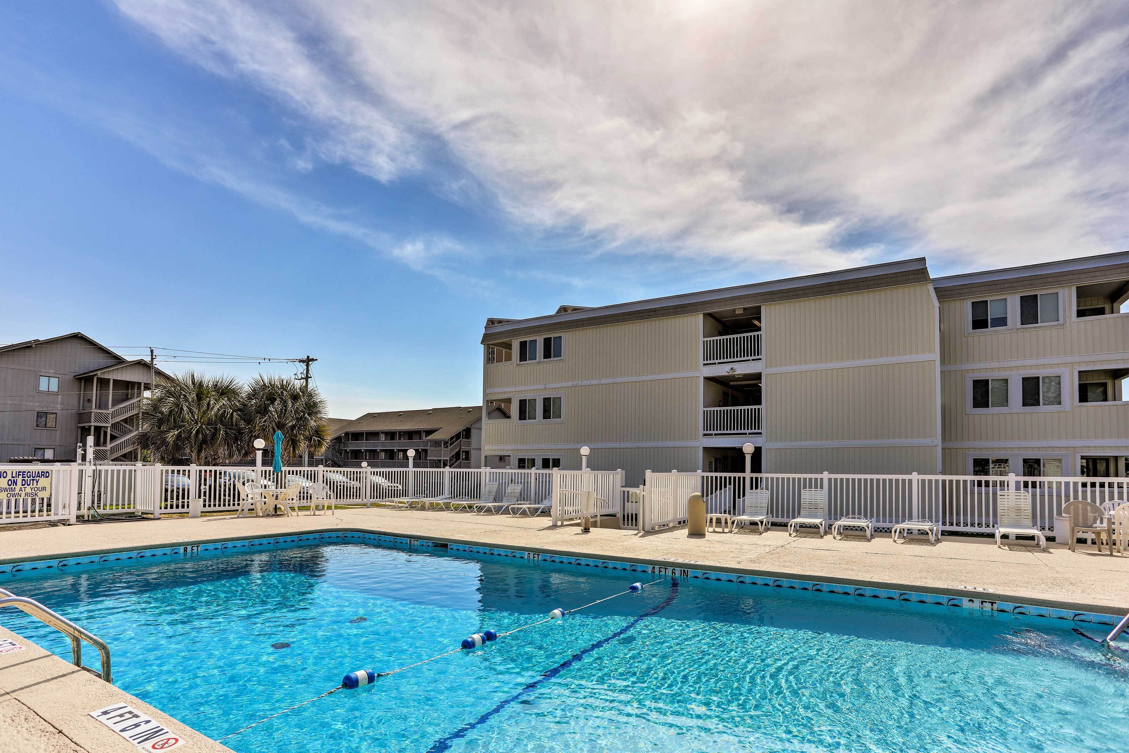 A swimming pool with a white fence, lounge chairs, and a multi-story building in the background.