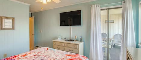 A bedroom with a bed, a dresser, a ceiling fan, a television, and a sliding glass door leading to a balcony with chairs.