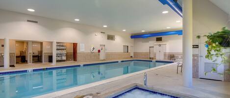 En indendørs swimmingpool med et afgrænset boblebad, fliserede vægge og et loft med indbygget belysning.