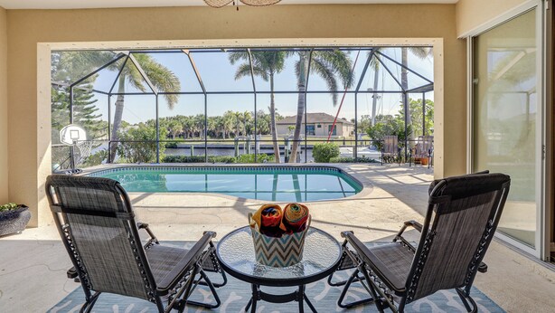 A poolside area with two chairs and a table, a ceiling fan, and a view of a swimming pool and palm trees. - Pet-friendly Waterfront Home w/ Pool! (Port Charlotte)