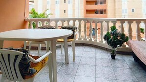 Un balcon avec une table et des chaises blanches, un pot de fleurs et une vue sur un immeuble à plusieurs étages.