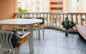 A balcony with a white table and chairs, a potted plant, and a view of a multi-story building.