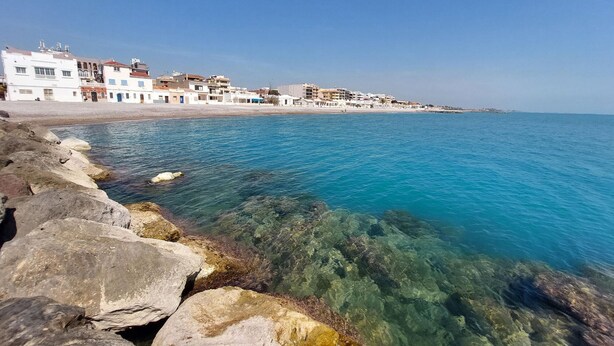 Une ville côtière avec des bâtiments qui bordent le littoral, une mer d’un bleu limpide et un premier plan rocheux.