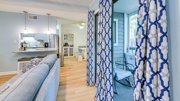 A modern living room with a sofa, a dining area with a kitchen in the background, and a patterned curtain. - Walkable Calabash Condo w/ Pool Access! (Calabash)