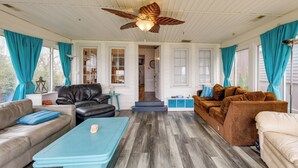 A spacious living room with a wooden floor, a ceiling fan, and large windows with teal curtains. - Lakefront Antioch Cottage w/ Dock & Gas Grill! (Antioch)