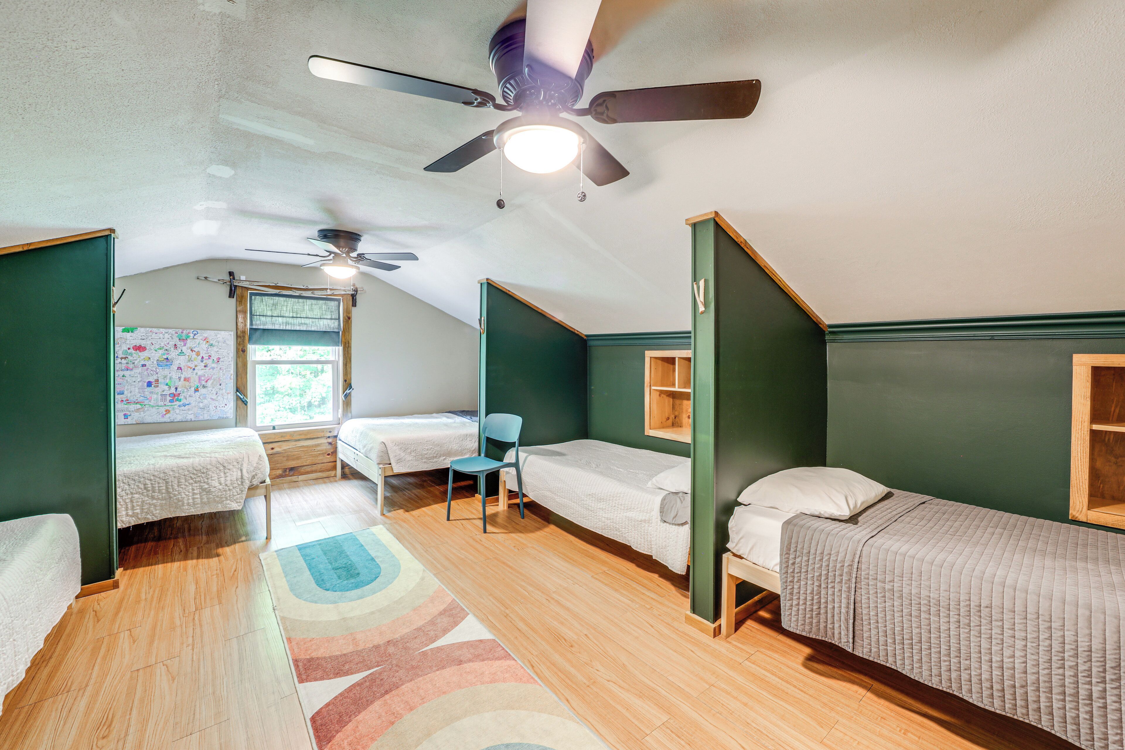 A dormitory room with two single beds, a ceiling fan, a window with a view, and a colorful rug.