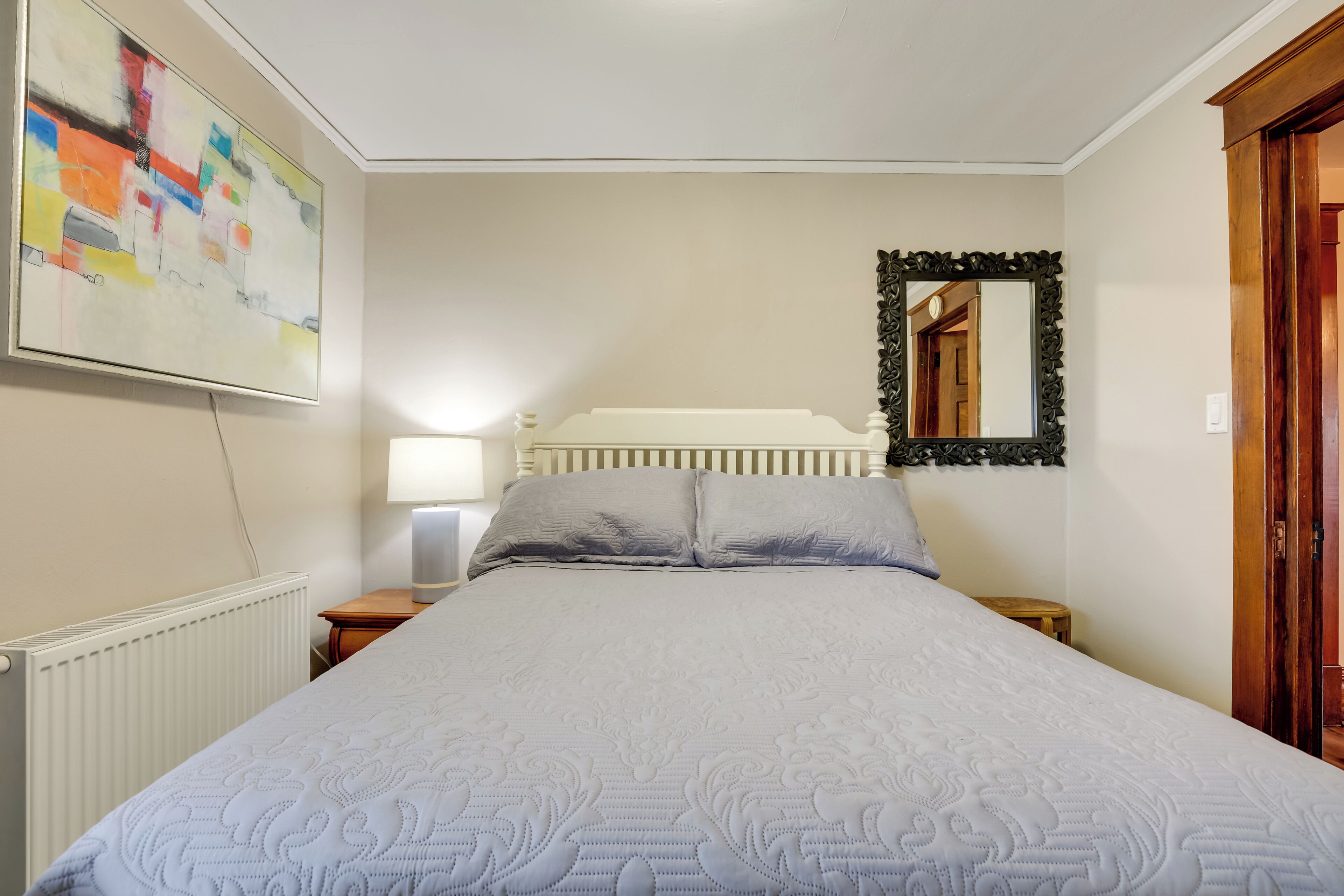A bedroom with a large bed, a bedside table with a lamp, a mirror, and a radiator.