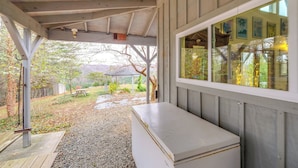 Une véranda avec un banc blanc, une terrasse en bois et une vue sur un jardin arboré, agrémenté d’un autre banc.