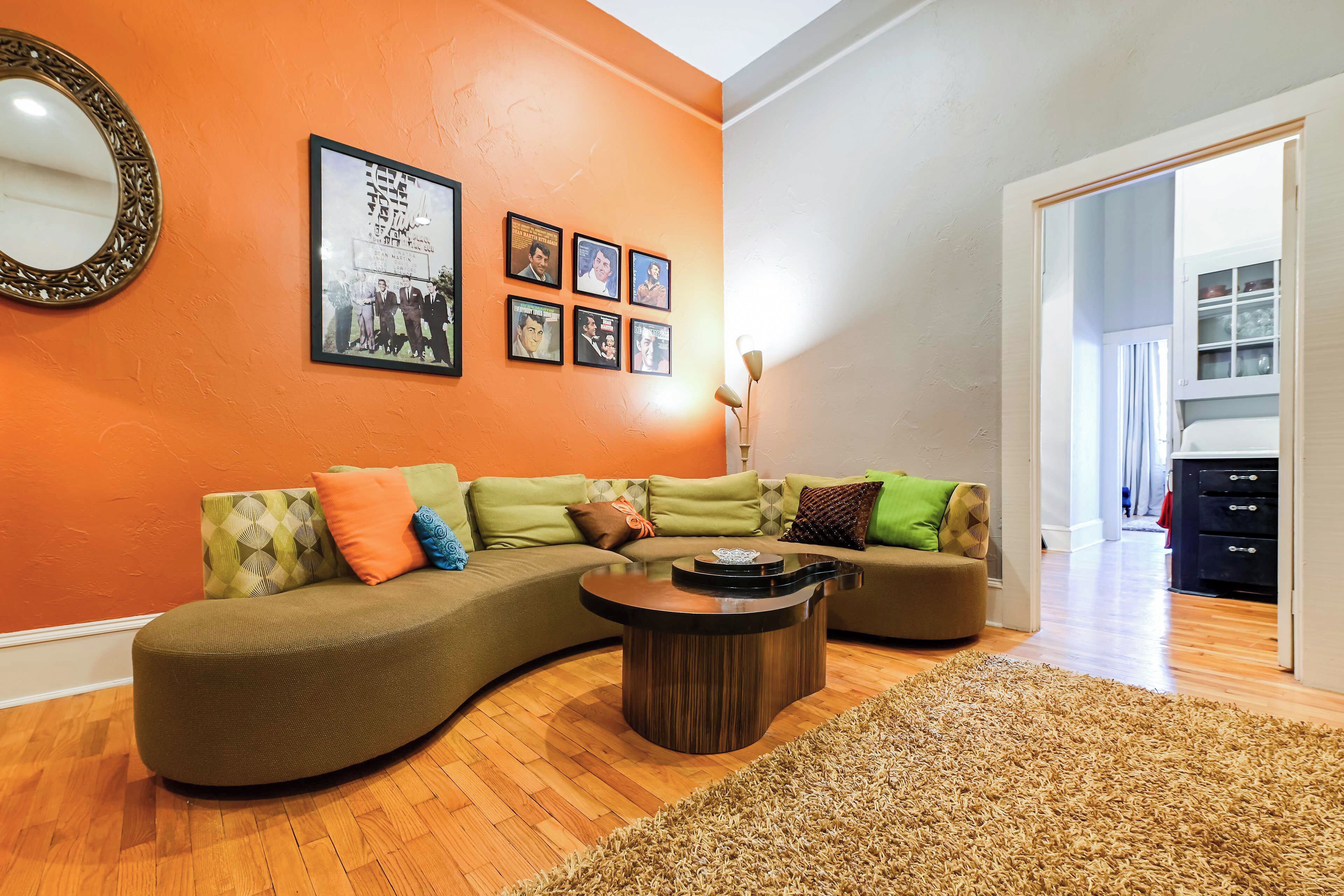 Una sala de estar con un sofá modular curvo, una mesa de centro redonda y una alfombra. Hay cuadros enmarcados en la pared y un espejo sobre el sofá.
