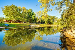 A serene lakeside setting with a dock, trees, and a building in the background. - Lakefront Nekoosa Home: Private Beach & Boat Dock! (Nekoosa)