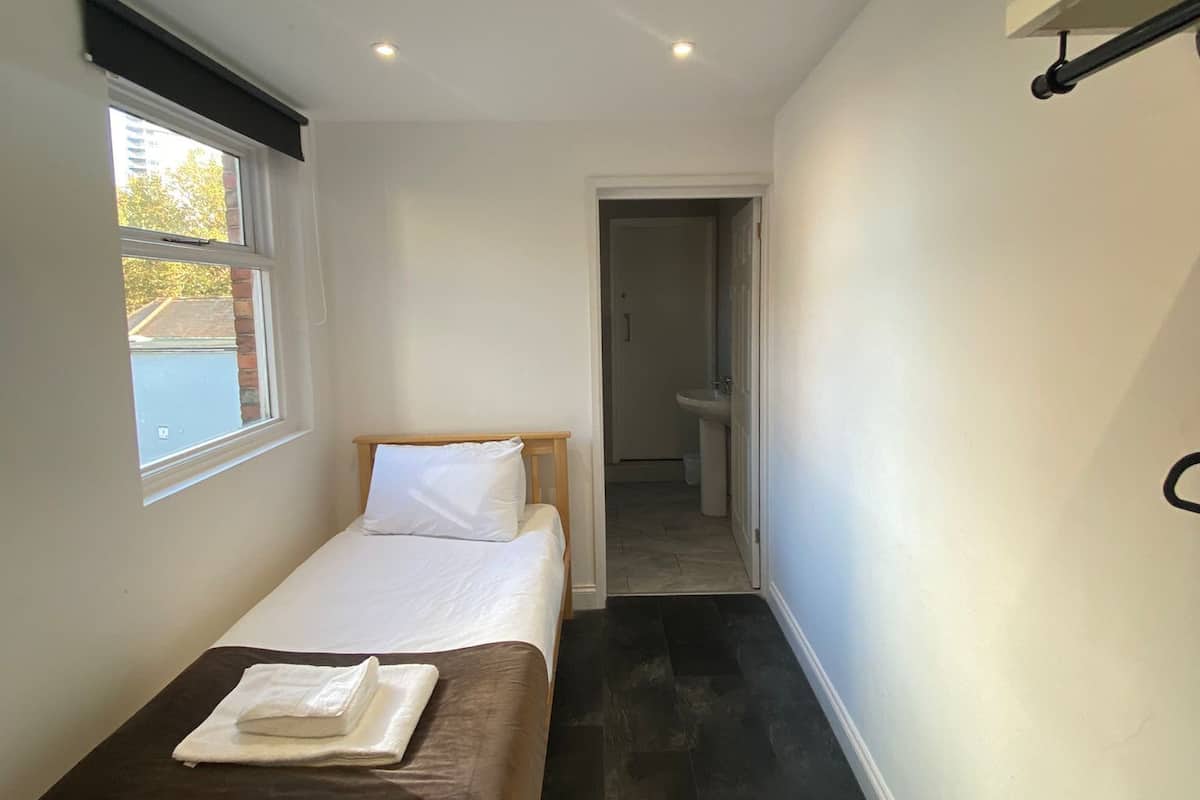 Een smalle, goed verlichte kamer met een eenpersoonsbed, een klein raam en een badkamer zichtbaar door een open deur.