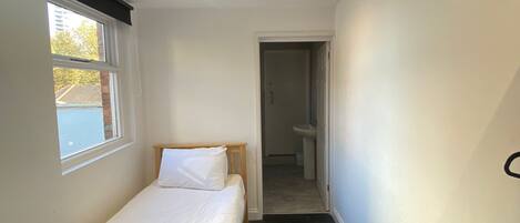 Una habitación pequeña y luminosa, con una cama individual, un ventanal y un baño visible a través de una puerta abierta.