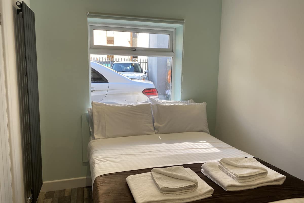 Een klein, net opgemaakt bed met witte lakens en twee opgevouwen handdoeken op een donkerbruine sprei. Een raam met uitzicht op geparkeerde auto's is zichtbaar.