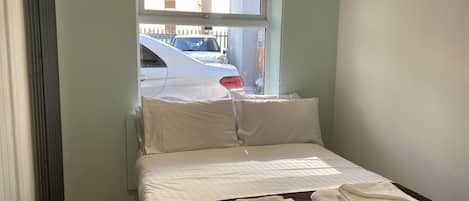 Een klein, net opgemaakt bed met witte lakens en twee opgevouwen handdoeken op een donkerbruine sprei. Een raam met uitzicht op geparkeerde auto's is zichtbaar.
