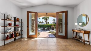 A room with a tiled floor, a wooden console table with a mirror, a shelf with decorative items, and a view of a patio with palm trees. - Mirador Desert Oasis – Mediterranean Escape (Palm Springs)