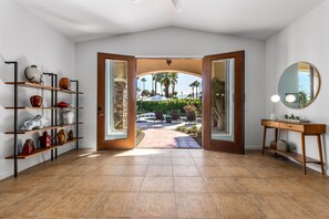 A room with a tiled floor, a wooden console table with a mirror, a shelf with decorative items, and a view of a patio with palm trees.