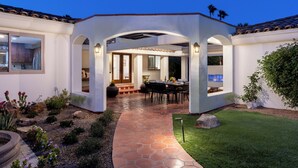 A covered patio with a dining area, a kitchen visible through an archway, and a well-maintained garden. - Mirador Desert Oasis – Mediterranean Escape (Palm Springs)
