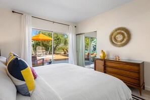 A bedroom with a large bed, a wooden dresser, a wall art piece, and a sliding glass door leading to an outdoor seating area. - Stargazer - Vibrant Mid-Century Retreat (Palm Springs)